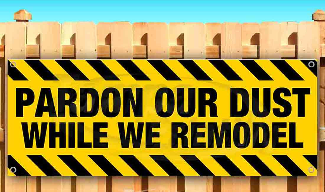 Yellow and black warning sign with 'Pardon Our Dust While We Remodel' text on a wooden fence.